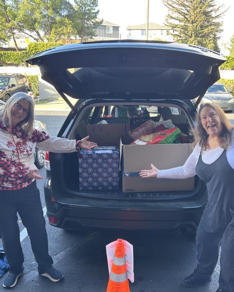 Two joyful woman with they're hands out in front of a car full of Christmas presents