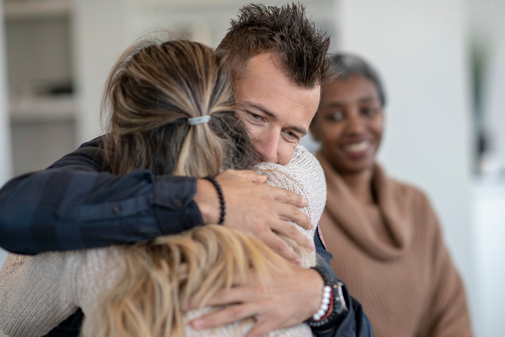 Man and woman hugging with another smiling woman looking on