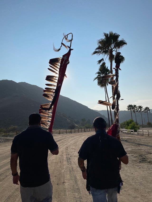 Two people walk down a wide dirt road at sunset, each carrying a tall ceremonial staff decorated with feathers.