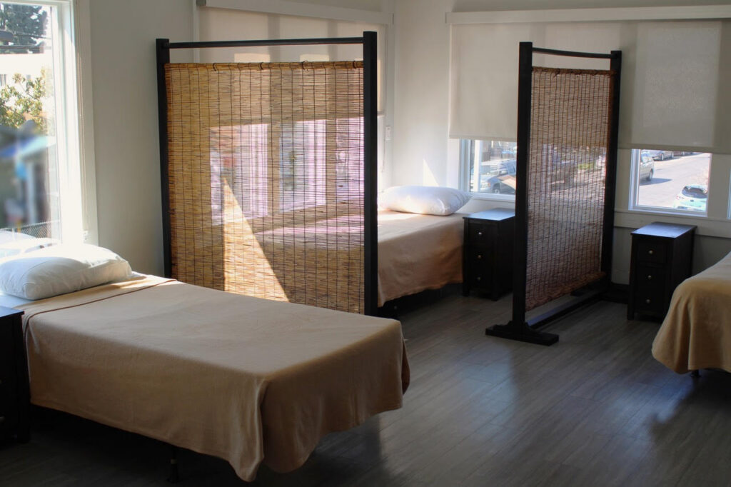 Sunlit room with three single beds, each separated by freestanding privacy dividers and small nightstands.